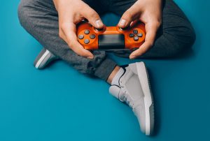 A young guy plays video games in his hands holding a red gamepad on a blue background, sitting in gray sneakers