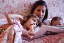 Mother reading to daughter (2-3 years) and baby (6-9 months) in bed