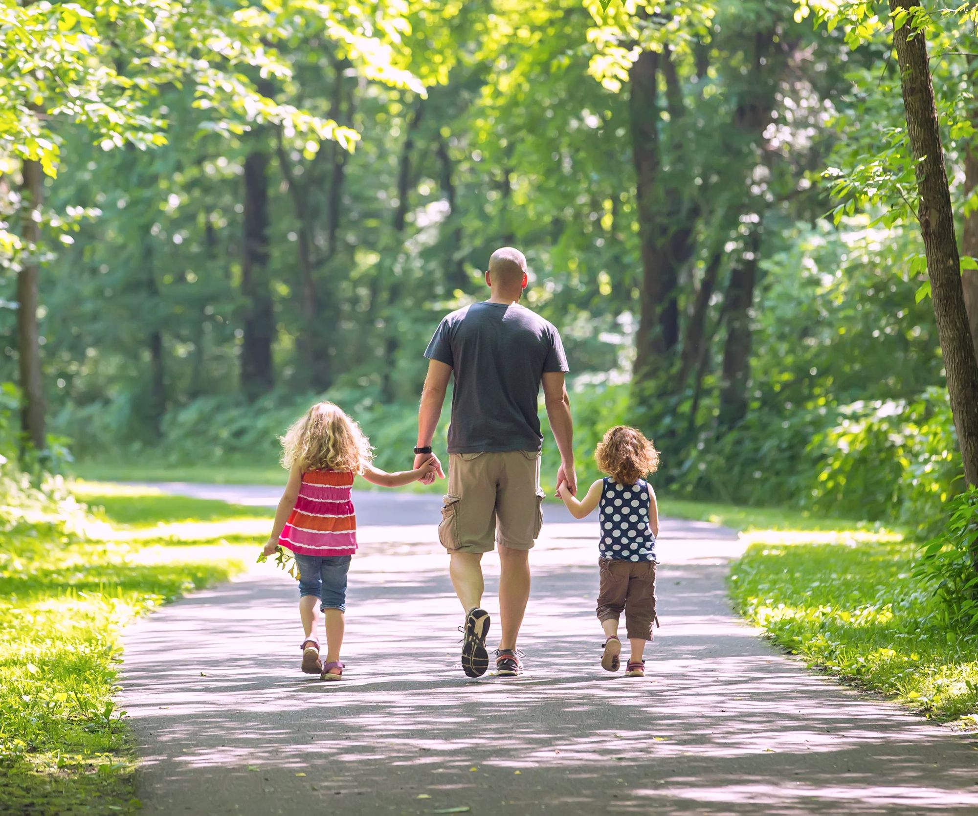 a father and his two young daughters going for a walk on a paved trail through the wooded park. 