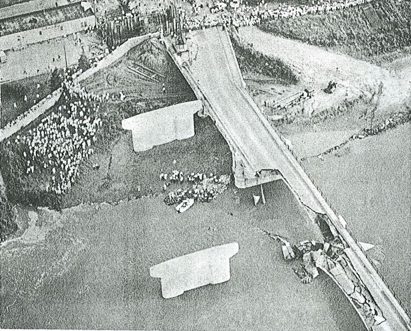 A black and white aerial image of the collapsed Melan bridge; the rescue workers can be seen in the river with a large crowd standing on the shore