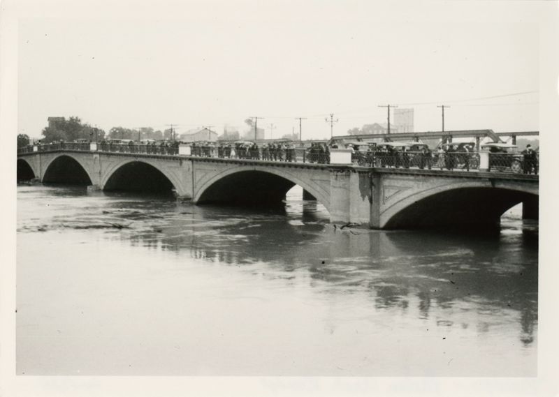 A black and white photo showing the Melan bridge during a flood; several people and cars are on the bridge