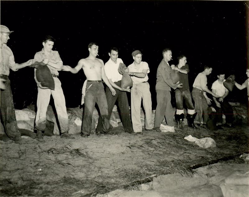 A black and white photo showing a line of men passing sandbags to each other during the 1951 flood