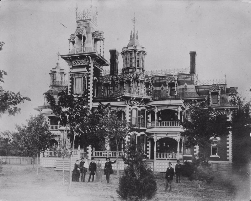 A black and white image of a elaborate three story mansion with several people on standing on the lawn 