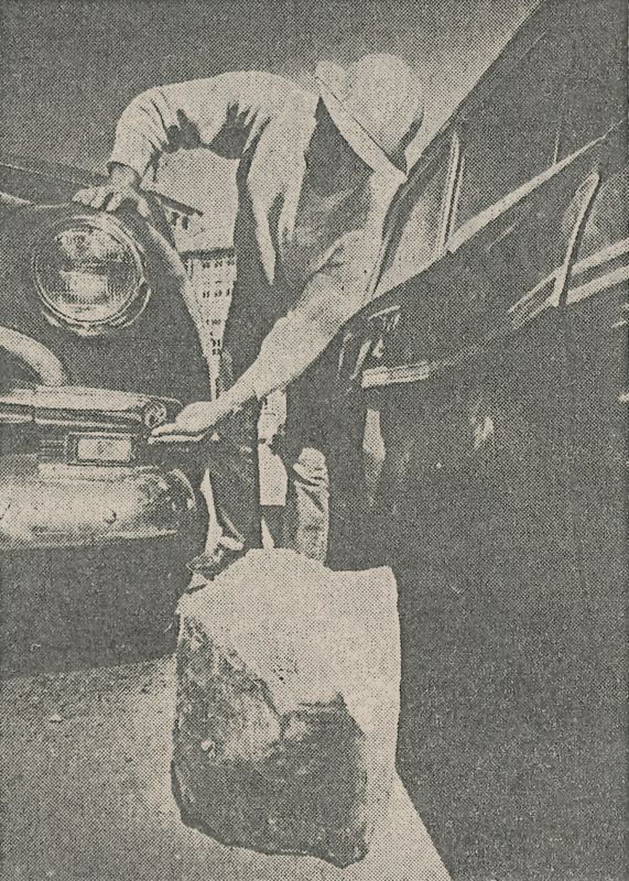 A black and white photo taken low between two cars with a large piece of stone in the foreground. A construction worker points to damage on one car