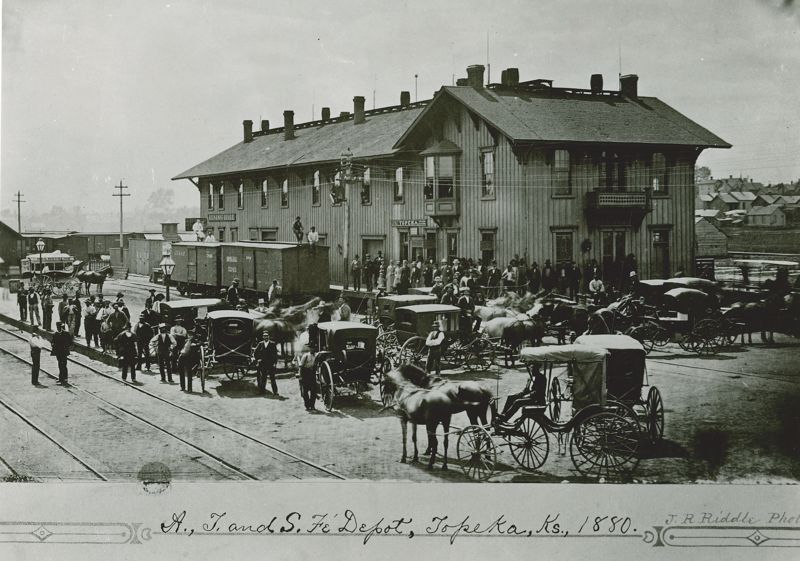 A black and white photo showing the Topeka Atchison, Topeka & Santa Fe depot in Topeka. The station is surrounded by buggies and carriages