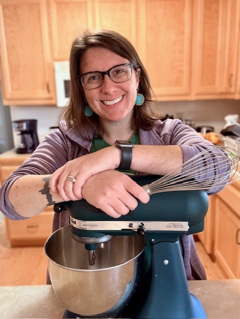 A color photograph of local history librarian, Katie Keckeisen; she is in her kitchen, arms crossed over her stand mixer, holding a whisk