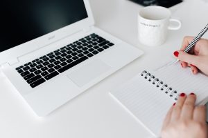 Full image of woman's hands writing in notepad with laptop and a cup of coffee near