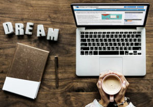 Condensed version of Featured image: Desktop with open laptop in front, writing notebook and DREAM written in lettering to left. A person's hands come in from the bottom of the frame and hold a cup of coffee at the edge of the laptop.