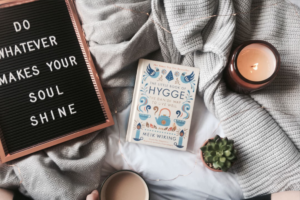 The Little Book of Hygge is surrounded by comfy blankets, a lit candle, succulents, and a sign that says, "Do whatever makes your soul shine." A person holds a cup of cocoa in the bottom of the frame.