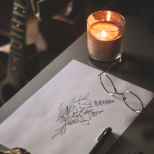 A table with a drawing of flowers, a pen, glasses, and a lit candle on it. There is a guitar in the background.