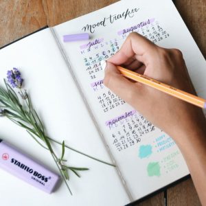 A woman's hand is updating a decorated bullet journal calendar with a pencil. Some lavender and a purple highlighter rest upon the opposite page.