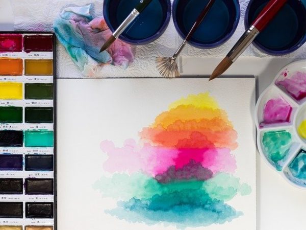 A white surface with a pan of watercolor paints, some vessels with colored water and paintbrushes, a palette with magenta, teal, and blue paints surrounding a piece of watercolor paper with layers of yellow, magenta and teal shapes painted upon it