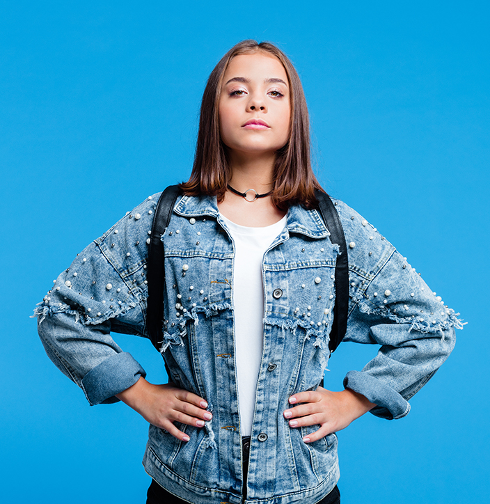 Confident female high school student wearing oversized denim jacket and white t-shirt, standing against blue background. Portrait of power girl.
