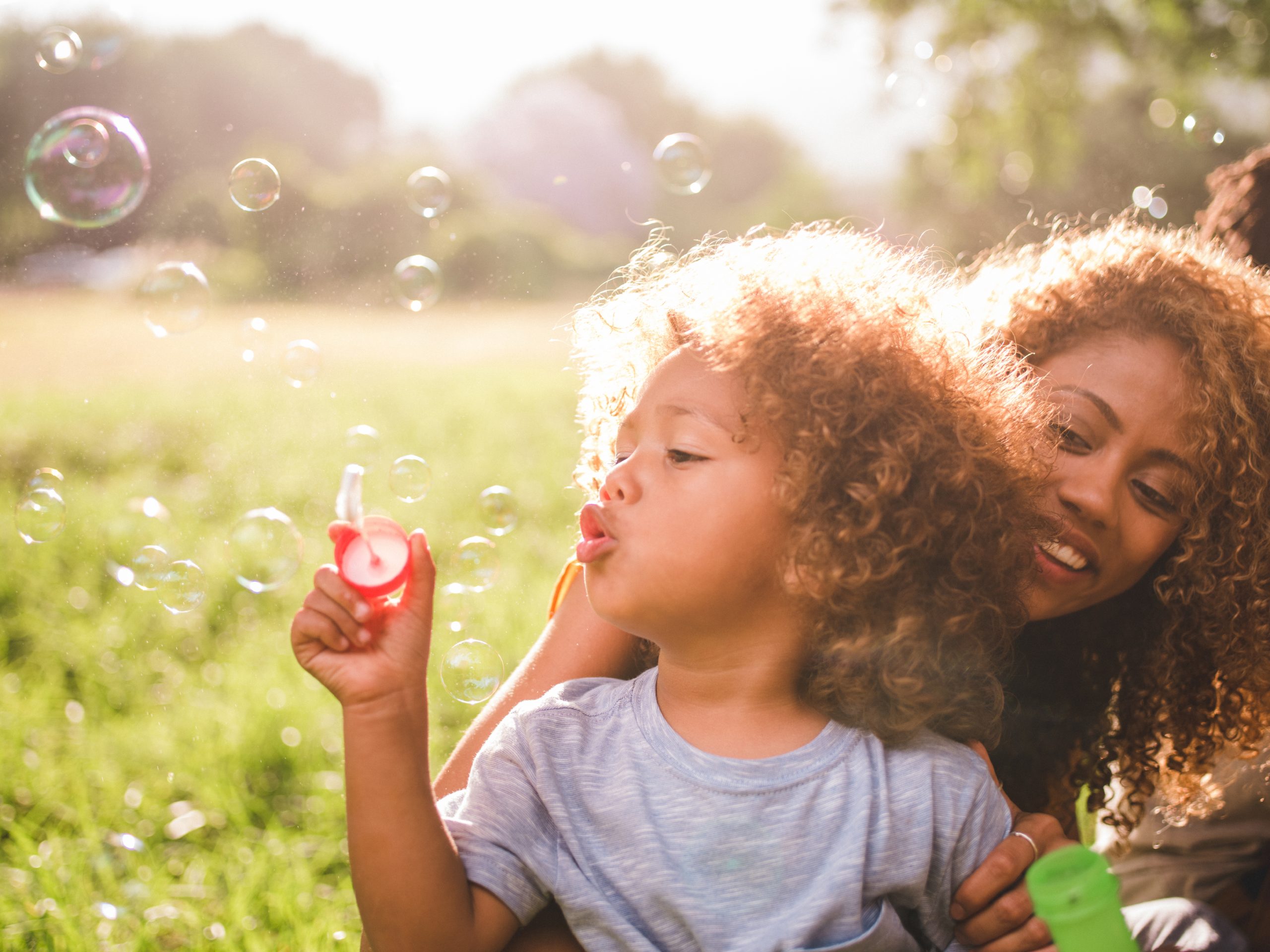 Young african-american mother and her cute curly haired baby boy blowing colourful bubbles on the meadow.
