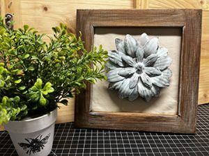 Gray ceramic floret placed in square, rustic wooden frame. Photographed near potted green plant.