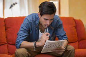 Young man sitting doing a crossword puzzle looking thoughtfully at a magazine with his pencil to his mouth as he tries to think of the answer to the clue